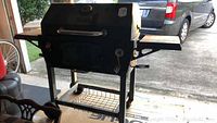 Full view of black Master Forge charcoal barbecue grill with hinged lid, glass window, side fold out shelves, and lower wire shelf on wheels located outdoors.