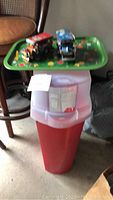 Tall plastic wrapping paper storage bin with red base and translucent top with 3 Christmas trays and 2 toy cars on top