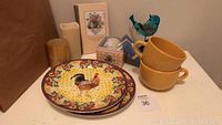 Photo showing two ceramic sandwich plates with colorful hen and sunflower design, two stacked yellow soup mugs, small blue bird figurine, gold candle holder, small floral memo book and a boxed tea cup set