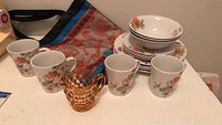 Floral patterned ceramic dinnerware set with bowls, plates, and four matched mugs, plus amber glass creamer on white table.