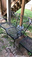Cast iron settee with mesh seat and decorative wrought iron scroll design under staircase on brick patio.