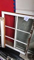 Large 6-pane window leaning against wall with visible chipping on white paint frame and clear glass panes.