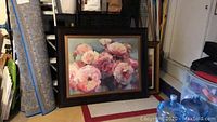 Framed floral painting resting on the floor surrounded by other framed artworks, rolled carpet, and plastic storage bins. The floral design predominantly features pink and peach blooms with green background. The frame is plastic with a wood colored finish.