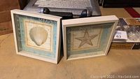 Two square sea-themed decorative wall frames placed side by side on floor, one featuring a seashell and the other a starfish.
