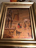 Close-up of antique or vintage framed tin painted picture showing a village scene with people, dogs, and windmill.