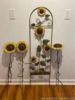 Wide shot showing the sunflower garden lattice with multiple yellow sunflowers, green metal leaves and stems, and the three individual metal sunflower stakes arranged next to it.