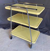 Front angled view of the vintage yellow metal kitchen cart with chrome frame and three shelves, shown on a light carpet.