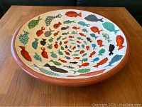Top angled view showing the large pottery plate with colorful fish painted in spiral pattern inside and terracotta rim.