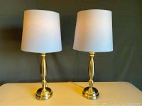Pair of brushed nickel table lamps with white tapered drum shades on a light table against a neutral background.