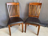 Two Ashley wooden dining chairs with vertical slat backs and black vinyl seats shown front-facing, positioned side by side on carpeted floor with a plain gray backdrop.