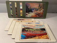 Eight cork placemats arranged fanned out showing their landscape art designs and varying colors typical of Group of Seven paintings.