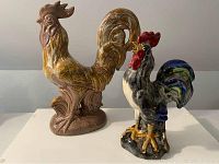 Pair of ceramic rooster figurines, larger brown with gold and smaller multicolored, both standing on a flat surface.