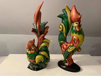 Pair of colorful ceramic rooster figurines displayed side by side on a white surface against a neutral wall. Both roosters have vibrant green, yellow, and orange hues with stylized feather detailing.