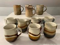 Front view of all eleven pottery mugs arranged with three large mugs on top shelf and eight smaller mugs on lower shelf, showing variety in size and glaze