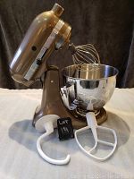 Side view of bronze KitchenAid Artisan tilt-head mixer with stainless steel bowl and attachments laid out.