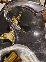 Side view of the chrome faucet showing curved spout and gold-tone handles on either side placed on black marble surface.