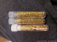 Three clear plastic vials filled with 24k gold flakes, lying horizontally on a dark fabric surface.
