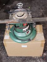 Photo shows a green Universal surface grinder with a 6-inch grinding wheel, mounted on a cardboard box. Visible metal adjustment handle and power cord are present.