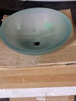 Photo showing the frosted light green glass sink placed on a cardboard surface, highlighting its round bowl shape and center drain hole.