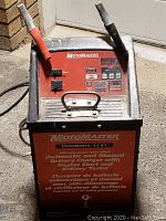 Photo of red and black MotoMaster battery charger with two large clamps and control panel with charging options and switches.