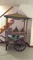 Front-side view of the vintage painted ice cream vendor cart showing palm tree and floral painted panels, metal containers on top, and peaked canopy roof.