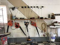 Photo showing two Black & Decker cordless leaf blowers and one Worx string trimmer hanging from wall-mounted pegboard. Batteries and chargers located on shelf above.