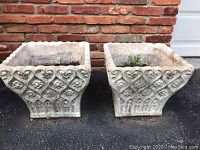 Two square concrete planters with ornate relief detail in front of brick wall