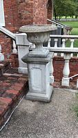 Full view of urn-style concrete planter sitting on a rectangular concrete pedestal next to brick steps and balustrade.