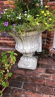 Full view of ornate concrete planter with multiple flowering plants and dirt inside, placed on brick surface.