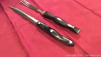 Pair of Kabar Cutco carving utensils on red fabric background showing serrated knife and fork with black handles