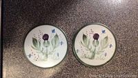 Pair of round ceramic trivets with hand-painted thistle designs, white base with green rims, placed on a speckled dark surface.