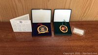 Two Christmas ornaments in open display boxes on a wooden surface with certificates and stand shown.