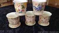 Five floral ceramic pots for African violets, arranged in two rows showing attached plates and varied flower patterns
