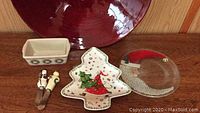 Photo showing the red bowl partially visible in background with 4 holiday-themed ceramic pieces in front: loaf pan, Christmas tree plate, frosting knives, crescent moon Santa plate.