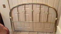 Photo of queen size brass headboard with curved top and vertical rods in polished brass finish leaning against a wall.