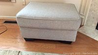 Front view of the light grey upholstered ottoman showing cushion top and espresso bun feet.
