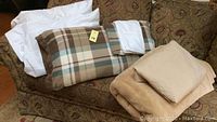 A beige fleece blanket with a beige pillowcase on top, two plaid fleece pillowcases with brown, cream, and light blue plaid design, and two white standard pillows arranged on a sofa.