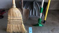 Photo of six long handled tools including broom, digging shovel, snow shovel, bulb changer and other tools lined up against a concrete wall on a floor.