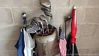 Full view of golf bag containing multiple golf clubs and umbrellas, positioned against concrete block wall.