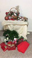 Full view of Christmas basket with ornaments, wreath on floor, red linens and other items on a beige cloth-draped table