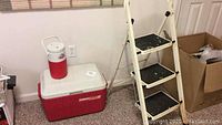 Wide view showing the red Coleman cooler with white lid, small insulated jug on top, and the cream-colored 3-step folding ladder with paint splatters beside it.
