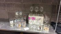 Wide view showing an assortment of multiple glasses placed on metal surface against cinderblock wall; includes various small and large glasses including some with designs and one box with wine glasses.