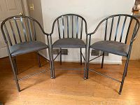 Three metal kitchen bar stools with gray padded seats, curved backs, and armrests shown together on wooden floor near a wall.