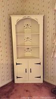 Front view of a white painted pine corner hutch cabinet, showing three open shelves with floral stencil designs on the back panels and two-door cabinet below with black metal hinges and handles.