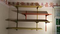 Photo showing adjustable shelving system mounted on wall with three floating shelves in gold, orange and avocado colors on metal racks.