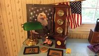 Photo showing nautical desk items including an eagle figurine on American flag base, framed Courage poster, wooden multiple dial barometer, small American flag, green vintage desk lamp, and various small decorative items.