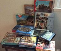 Overall view of 11 jigsaw puzzles stacked on table against beige wall. Various puzzle brands and piece counts seen. Puzzle box art shows castles, villages, boats, and autumn trees.