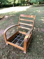 Side angle photo of the wooden Heywood Wakefield armchair frame outdoors on grass, showing slatted backrest and curved armrests with focus on chew marks on one armrest.