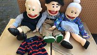 Photo of three large puppets: elderly man, elderly woman, and younger man sitting on a tabletop with one striped clothing item and metal rods visible
