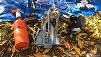 Overview showing galvanized boat anchor, Olin marine signal kit in orange case, air horn foghorn, Instant Power device, rope handle, and marine buoy on a blue tarp with fallen leaves
